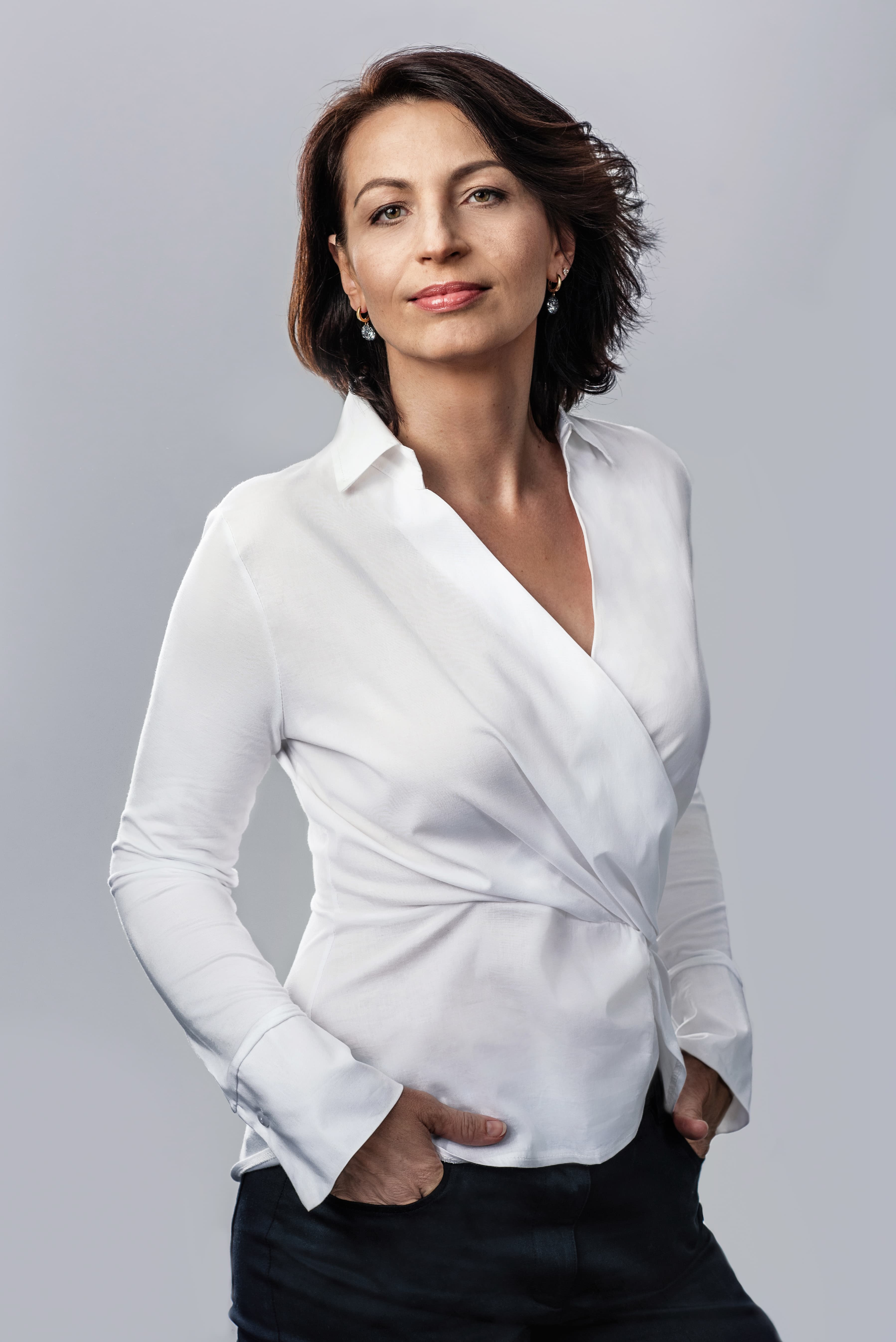 Woman with short brown hair wearing a white wrap blouse and dark pants, grey background.