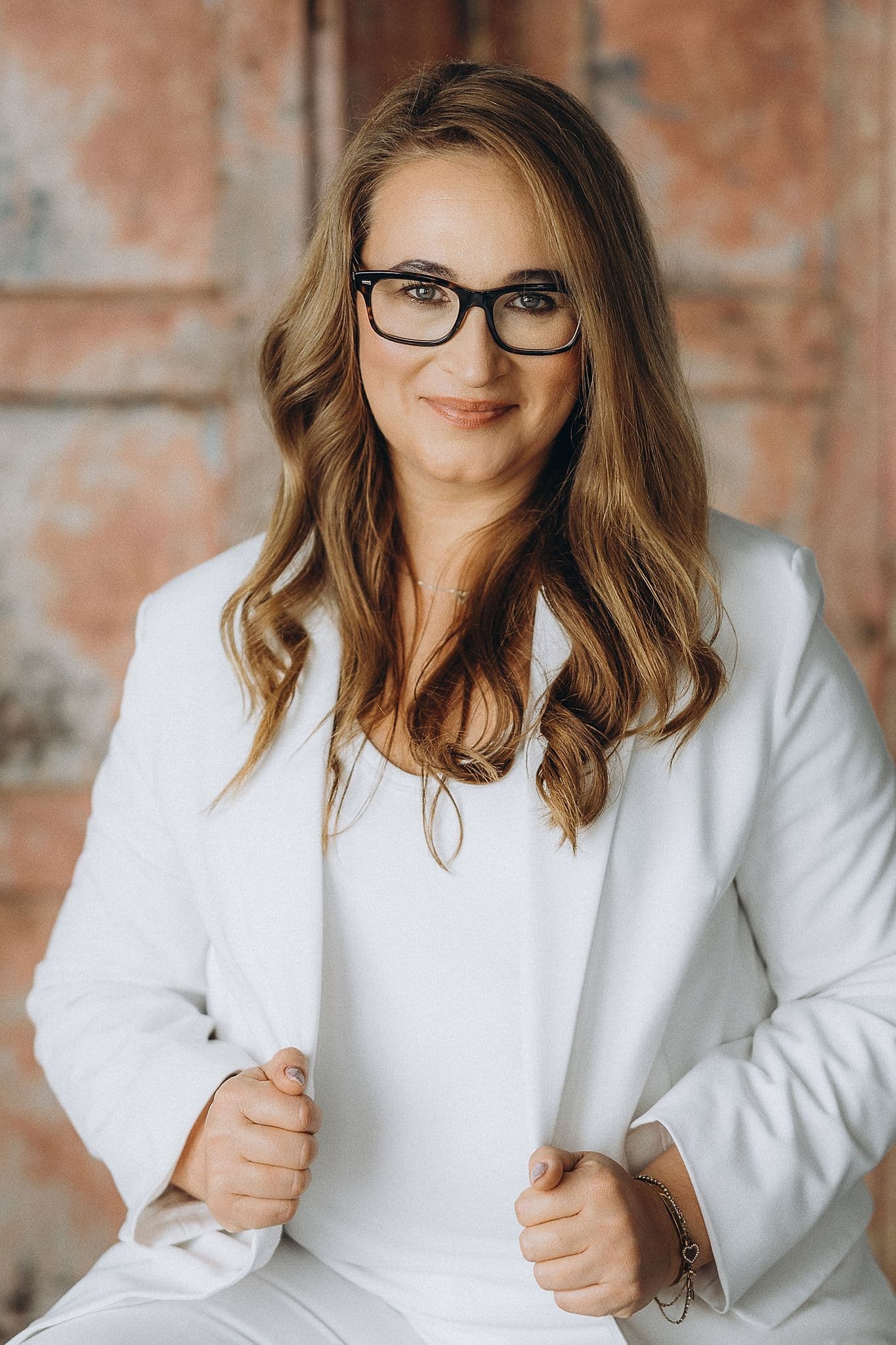 Woman with wavy brown hair and black glasses wearing a white blazer against textured background.