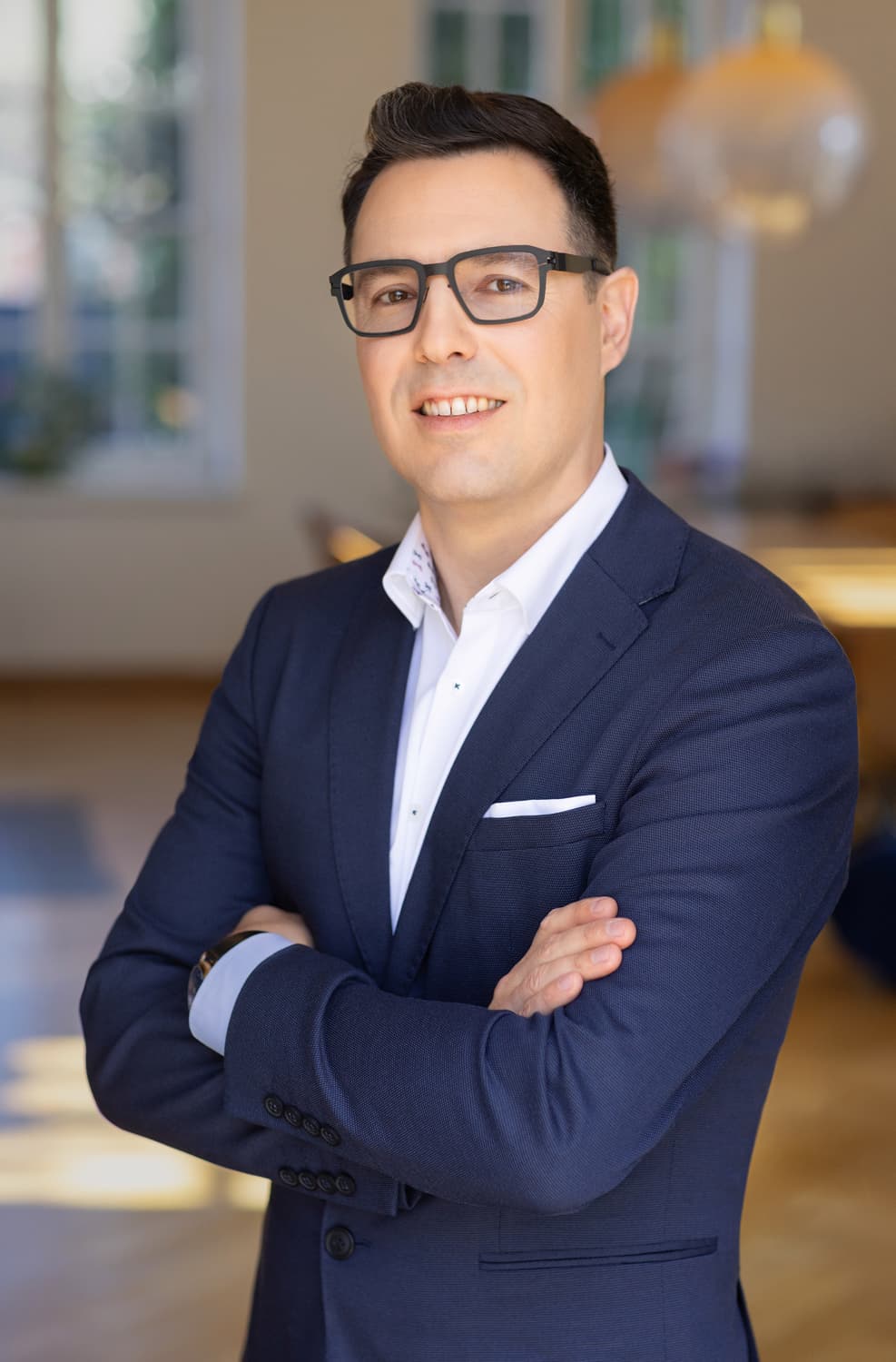 Smiling man in a navy blazer and glasses with arms crossed in a bright office.